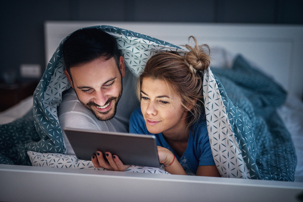 entzückendes junges entspanntes Paar, das unter einer Decke auf seinem schlechten Bett liegt und Nachrichten auf dem Tisch liest. - Foto, Bild