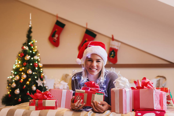 jovem mulher feliz vestindo chapéu de Papai Noel comemorando o Natal em casa com presentes na cama
 - Foto, Imagem