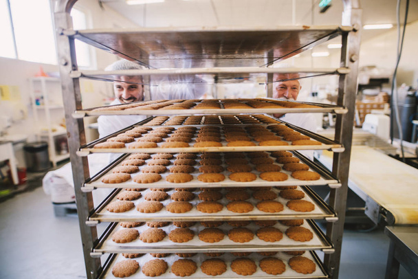 Foto van twee mannelijke bakkers in het witte uniform duwen elkaar rekken vol met verse gebakken koekjes. - Foto, afbeelding