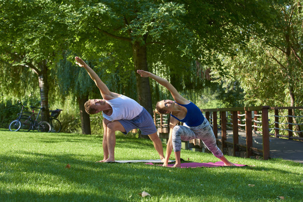 Full length shot of young man and woman training yoga together in the park. Couple doing yoga outdoor. - Foto, imagen
