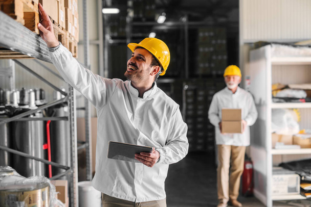 Picture of smiling young man warehouse worker counting boxes in warehouse. Holding digital tablet in his hands. - Photo, Image