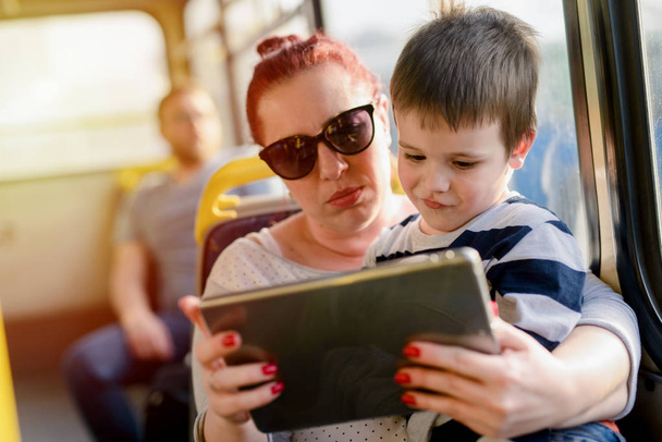 Nainen tilalla pieni lapsi poika ja osoittaa tabletti pc matkustaessaan bussilla
 - Valokuva, kuva