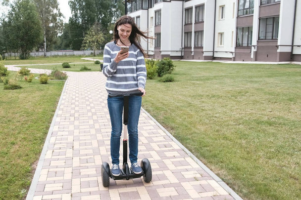 Beautiful brunette woman is rolling on gyro scooter near the home and looking at mobile phone. - Foto, Bild