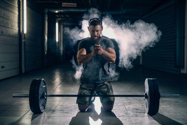 Muscular attractive caucasian bearded man smiling, kneeling, clapping hands with chalk powder, preparing to lift barbell in underground hallway. - Photo, Image