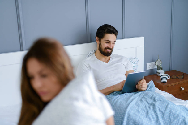 young sad woman sitting on bed, boyfriend with tablet pc in hands on background - Foto, imagen