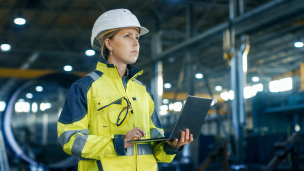 Female Industrial Engineer in the Hard Hat Uses Laptop Computer while Standing in the Heavy Industry Manufacturing Factory. In the Background Various Metalwork Project Parts Lying - Foto, afbeelding