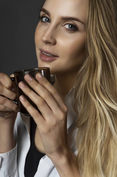 A beautiful smiling pregnant young female blonde wearing a black maternity underwear and a white unbuttoned shirt, tied on her stomach, holds a cup of hot drink in her hands. Copy space. Close up. - Fotó, kép
