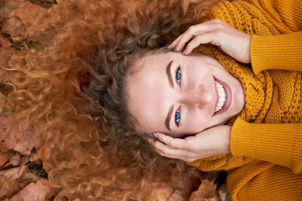 Redhead curly cheerful cute girl liying in the autumn leaves, holding face and smiling. Autumn portrait. Top view. - Foto, imagen