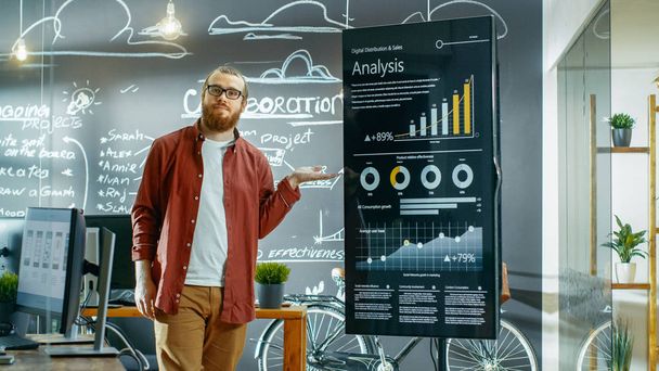 Male Developer Looks at Interactive Whiteboard with Charts, Graphs and Growth Statistics. He Points at Display with Presenting Gesture and Smile. In the Background Creative Office with Chalkboard Wall - Zdjęcie, obraz