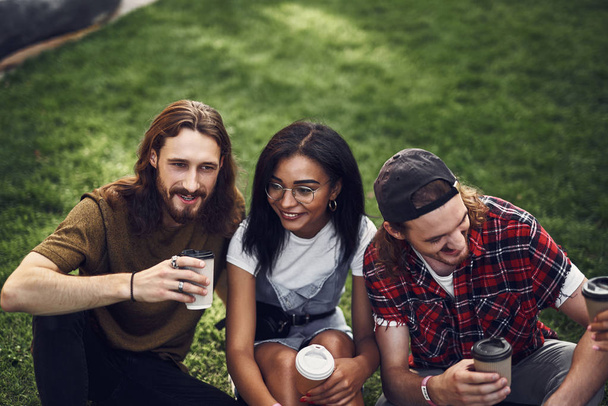 Tre giovani amici che si godono il caffè seduti sull'erba insieme
 - Foto, immagini