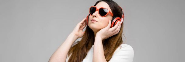 Portrait of emotional beautiful plus size model in headphones and sunglasses standing in studio looking away listening to music - Photo, Image
