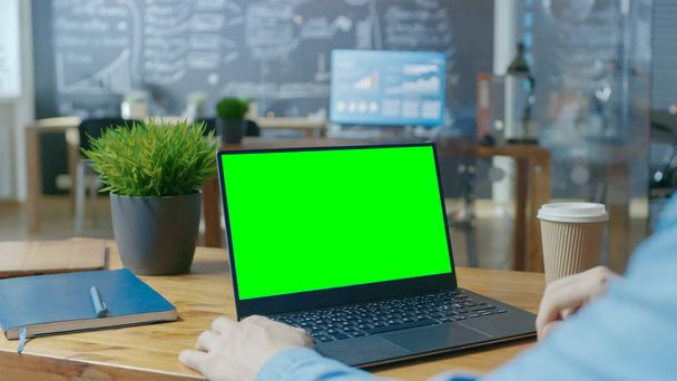 Male Office Worker at His Desk Works on a Laptop with Mock-up Green Screen. Over the Shoulder Close-up Footage. In the Background Creative Office. - Foto, afbeelding