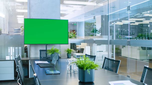 moderner, leerer Besprechungsraum mit großem Konferenztisch mit verschiedenen Dokumenten und Laptops darauf, an der Wand großer Fernseher mit grünem Chroma-Schlüsselbildschirm. - Foto, Bild