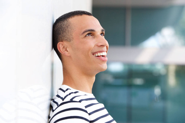 Profile portrait of happy young man leaning against white wall  - 写真・画像