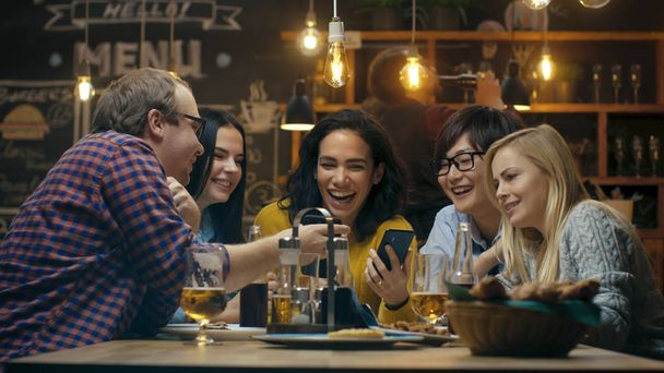 Krásné hispánské žena ukazuje zajímavé věci na její Smartphone na její přátele, zatímco oni mají dobrý čas v baru. Směje se, vtip, pít v podniku stylového baru Hipster. - Fotografie, Obrázek