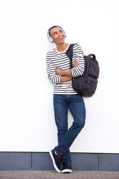 Ganzkörperporträt eines glücklichen jungen Mannes, der sich mit Tasche und Kopfhörer an eine weiße Wand lehnt - Foto, Bild