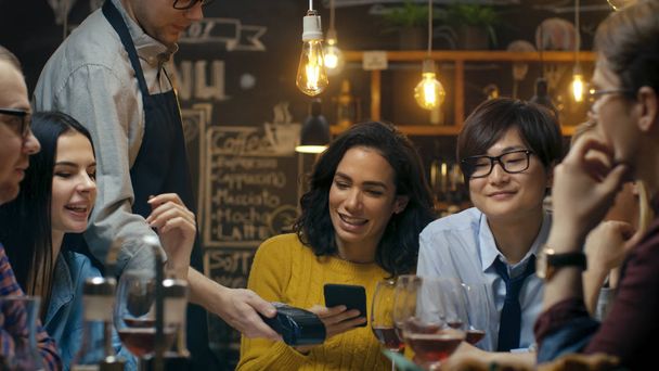 In the Bar Waiter Holds Credit Card Payment Machine and Beautiful Woman Pays for Her Order with Contactless Mobile Phone Payments System. She Has Good Time with Her Friends. - 写真・画像