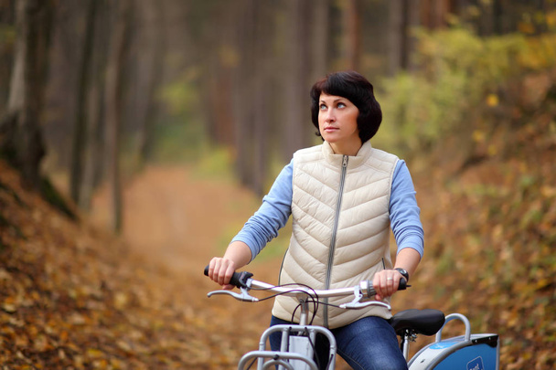 The girl on a bike from the rental in the forest. Beautiful young woman posing for the camera in the autumn forest.  - Фото, изображение