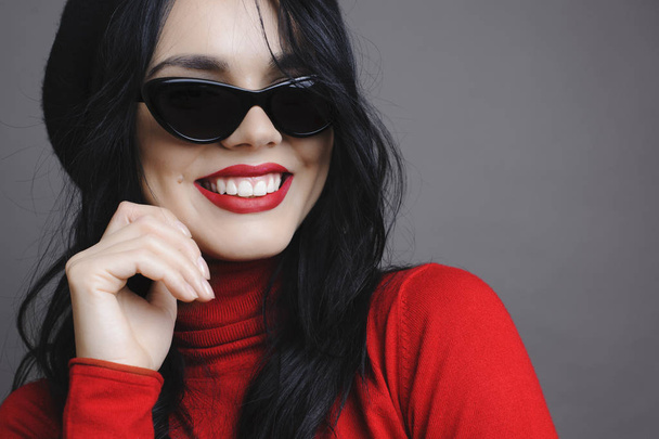 Lovely young woman in stylish sunglasses smiling and looking away while standing on gray background - Foto, imagen