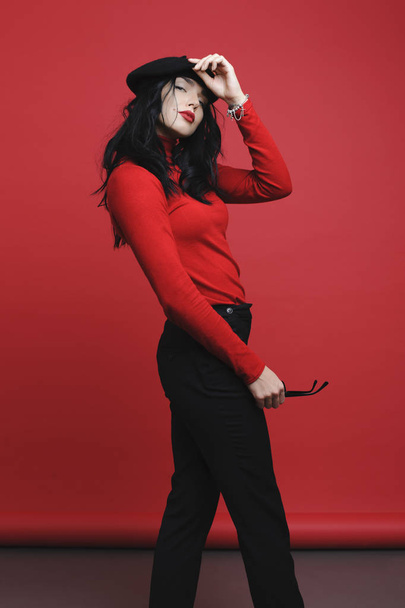 Side view of attractive young lady in trendy outfit touching beret and looking at camera while standing on bright red background - Foto, afbeelding