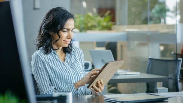Beautiful Young Hispanic Woman sits at Her Desk and Uses Tablet Computer. In the Background Busy Office with Working Colleagues. - 写真・画像