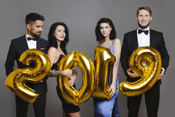 attraktive Männer in eleganten Anzügen und schöne Frauen in Abendkleidern mit goldenen Luftballons, die auf grauem Hintergrund die Schrift für 2019 bilden - Foto, Bild