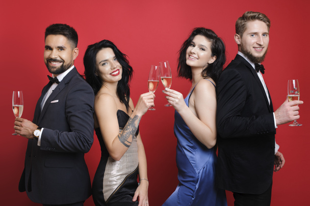 Zijaanzicht van knappe jongens in pakken en aantrekkelijke vrouwen in prachtige jurken holding glazen champagne en glimlachen terwijl staande op heldere rode achtergrond - Foto, afbeelding
