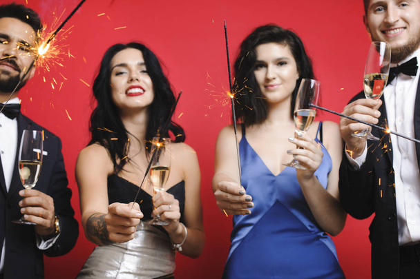 Group of young friends in elegant outfits holding glasses of champagne and burning sparklers while standing on red background - Photo, Image