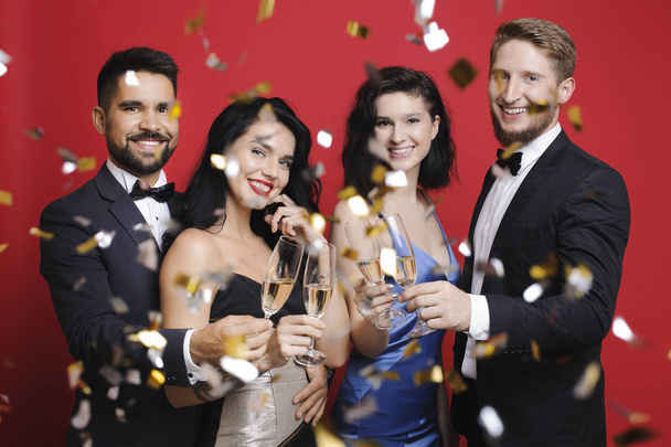 Two charming couples in evening outfits smiling and holding glasses of champagne while standing under falling confetti on vibrant red background - Photo, Image