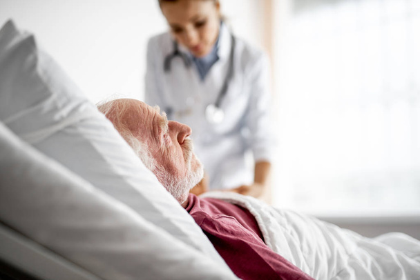Παλιά ο άνθρωπος αναπαύεται στο δωμάτιο του νοσοκομείου ενώ ο γιατρός ελέγχει την κατάστασή του - Φωτογραφία, εικόνα