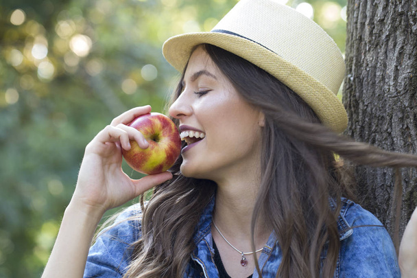 Beautiful young woman with a hat enjoying nature and eating an apple - Foto, afbeelding