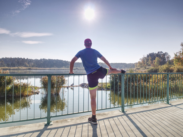 Мужчина разминает ноги перед тем, как выйти наружу в солнечное утро. Разогреваю мышцы. Упражнения на открытом воздухе на озерном мосту
. - Фото, изображение
