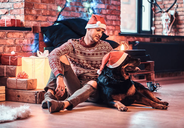 Молодий чоловік сидить зі своїм чистим ротвейлером у прикрашеній вітальні в різдвяний час
. - Фото, зображення