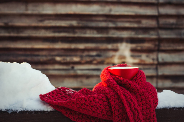 Ερυθρό κύπελλο με ζεστό τσάι και το κόκκινο φουλάρι στο χιονισμένο ξύλινα φόντο. Έννοια του χειμώνα. Ηλιόλουστη μέρα. Χειμερινές διακοπές. - Φωτογραφία, εικόνα