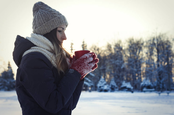 Una joven con guantes rojos sostiene una taza roja con té caliente en el fondo del paisaje de invierno al atardecer. Concepto de invierno
. - Foto, Imagen