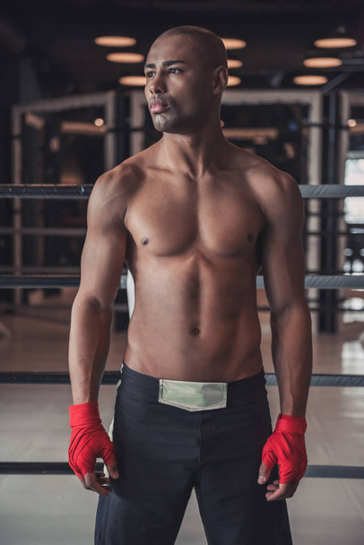 Afro American boxer, his hands wrapped in red bandage, is looking away while standing in pose in gym - Foto, Bild
