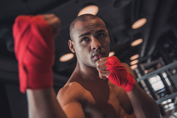 Afro American boxer, his hands wrapped in red bandage, is standing in boxer pose in gym - Photo, Image