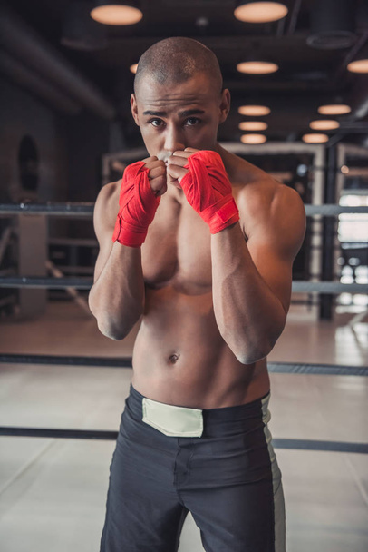 Афро-американский боксер, его руки завернуты в красную повязку, стоит в боксерской позе в спортзале и смотрит в камеру
 - Фото, изображение