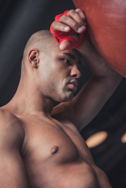 Tired Afro American boxer, his hands wrapped in red bandage, is leaning on the punching bag while training - Foto, immagini
