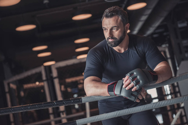 Привлекательный мускулистый боксер в боксёрских перчатках опирается на боксёрское кольцо и отворачивается во время тренировки в спортзале
 - Фото, изображение
