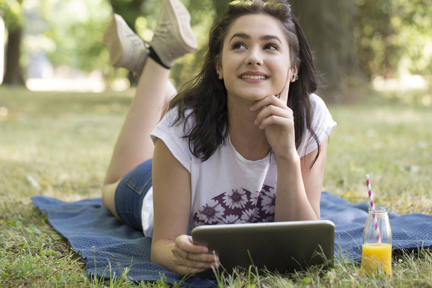 Mooi meisje, tiener, liggend op het gras en houden van een tablet. Ze is nadenkend - Foto, afbeelding