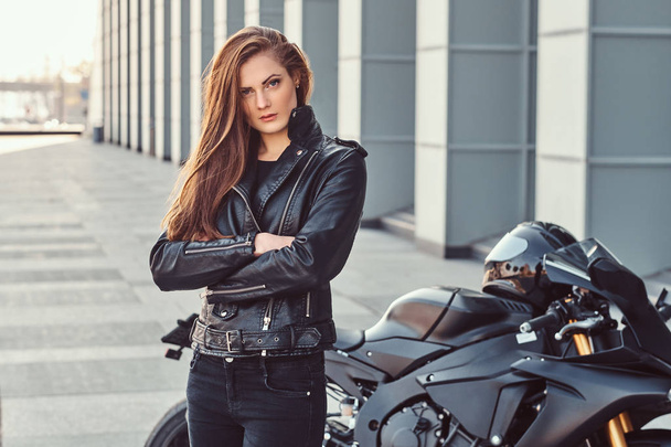彼女の腕に自信を持ってバイクに乗る女の子は建物の外の彼女のスーパー バイクの横に交差. - 写真・画像
