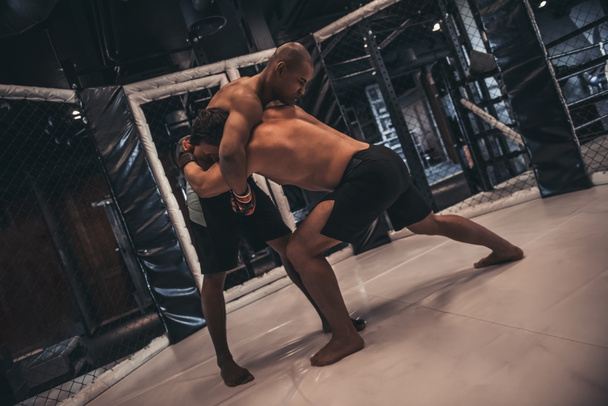 Two men in boxing gloves and shorts are fighting in cage using grappling technique - Photo, Image