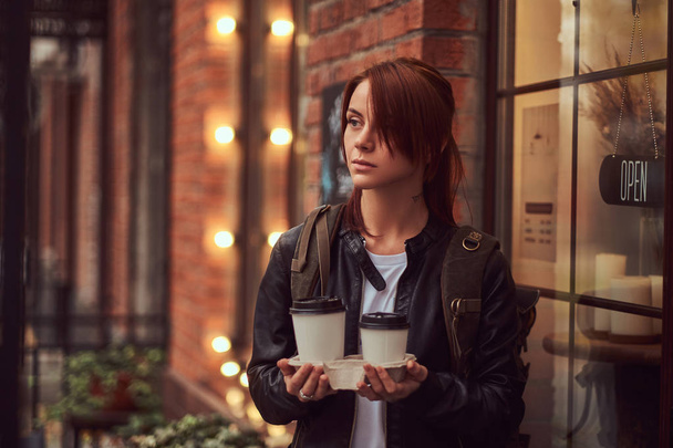 Urocza dziewczyna na sobie skórzaną kurtkę z plecakiem trzymając kubek z kawa na wynos poza w pobliżu kawiarni. - Zdjęcie, obraz