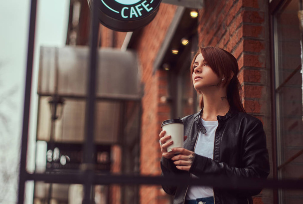 Viehättävä tyttö yllään nahkatakki reppu tilalla kuppi takeaway kahvia ulkopuolella kahvila
. - Valokuva, kuva