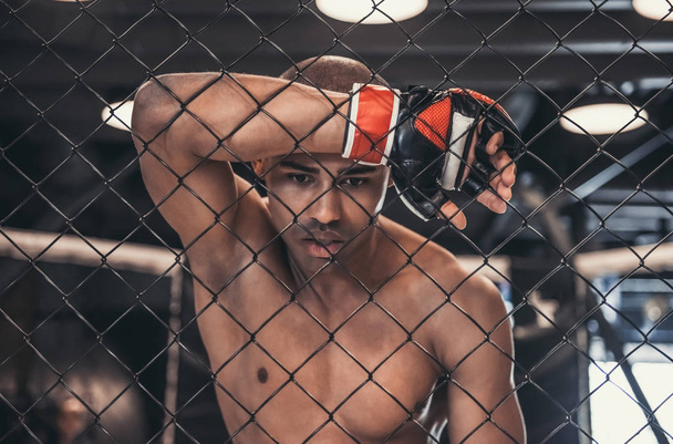 Tired Afro American fighter in gloves is leaning on the net of the cage - Foto, immagini