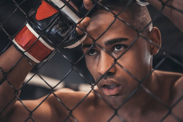 Cansado luchador afroamericano en guantes se apoya en la red de la jaula
 - Foto, imagen