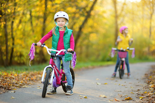 Cute little sisters riding bikes in a city park on sunny autumn day. Active family leisure with kids. Children wearing safety hemet while riding a bicycle. - Foto, immagini