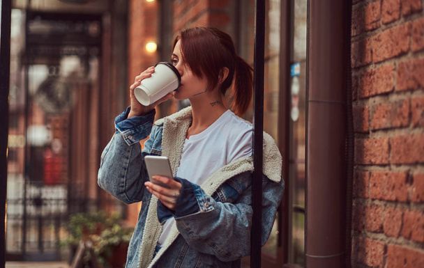 A beautiful tattooed girl wearing a denim coat drinks takeaway coffee holding a smartphone outside the cafe. - Photo, image