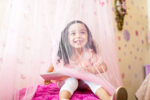Criança feliz do sexo feminino com tablet digital olhando através de cortina no quarto
 - Foto, Imagem